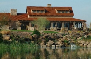 A large, two-story house with a red-tiled roof, a stone chimney, and a stone porch with columns. In front of the house is a rocky landscape with a small pond and a waterfall.