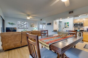 A modern living room with a brown sofa, wooden dining table, and chairs. There is a TV, a window with a view, and a kitchen area with cabinets and appliances.