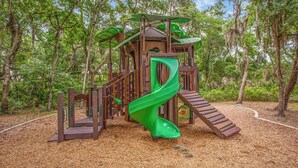 Una estructura de juego de madera con un tobogán verde y elementos para escalar, rodeada de árboles.