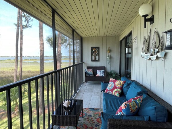 A covered porch with a view of a water body and trees, furnished with a blue sofa and a wicker chair.