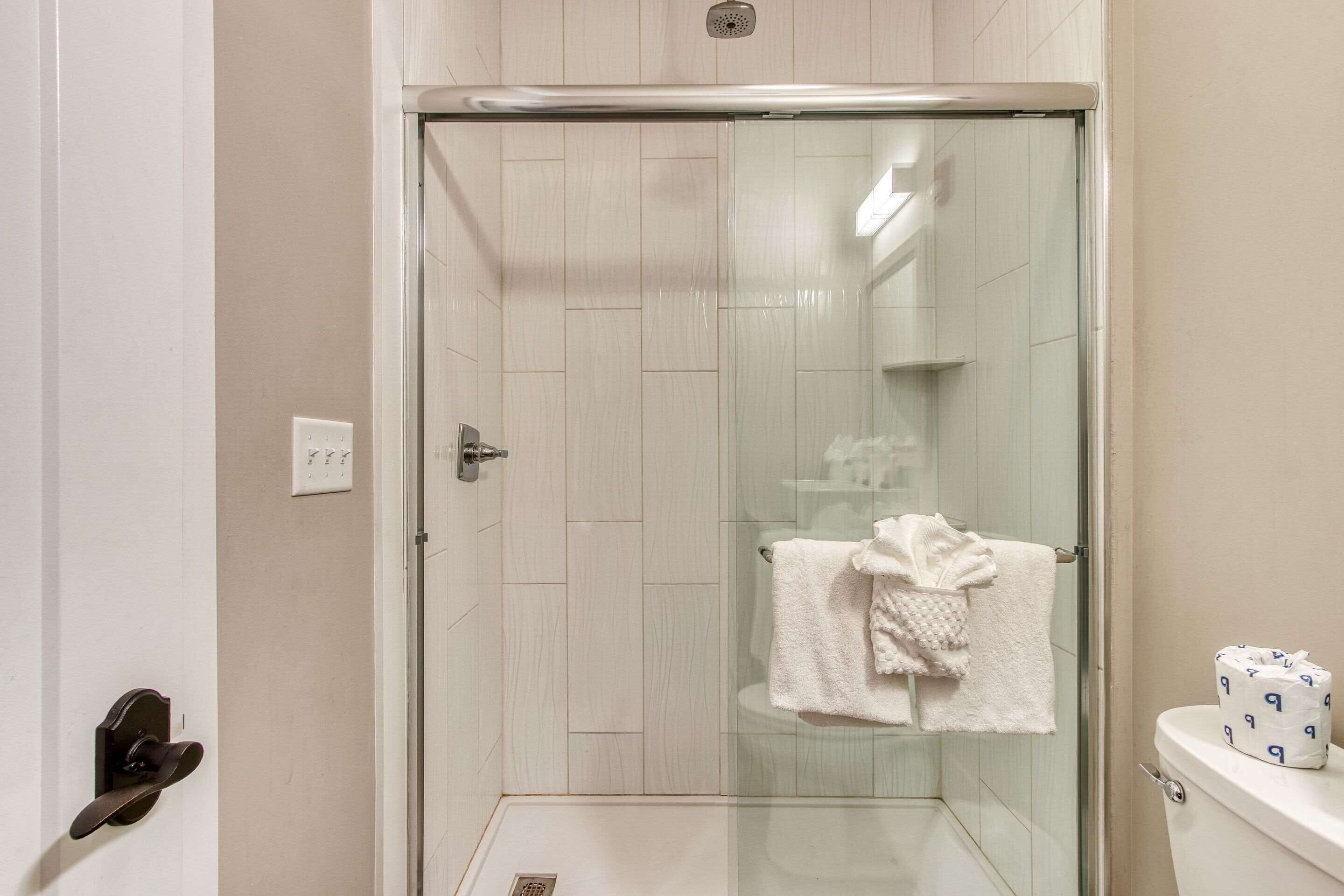 A glass-enclosed shower with a white tiled wall, a white towel on the wall, and a toilet with a paper towel holder.