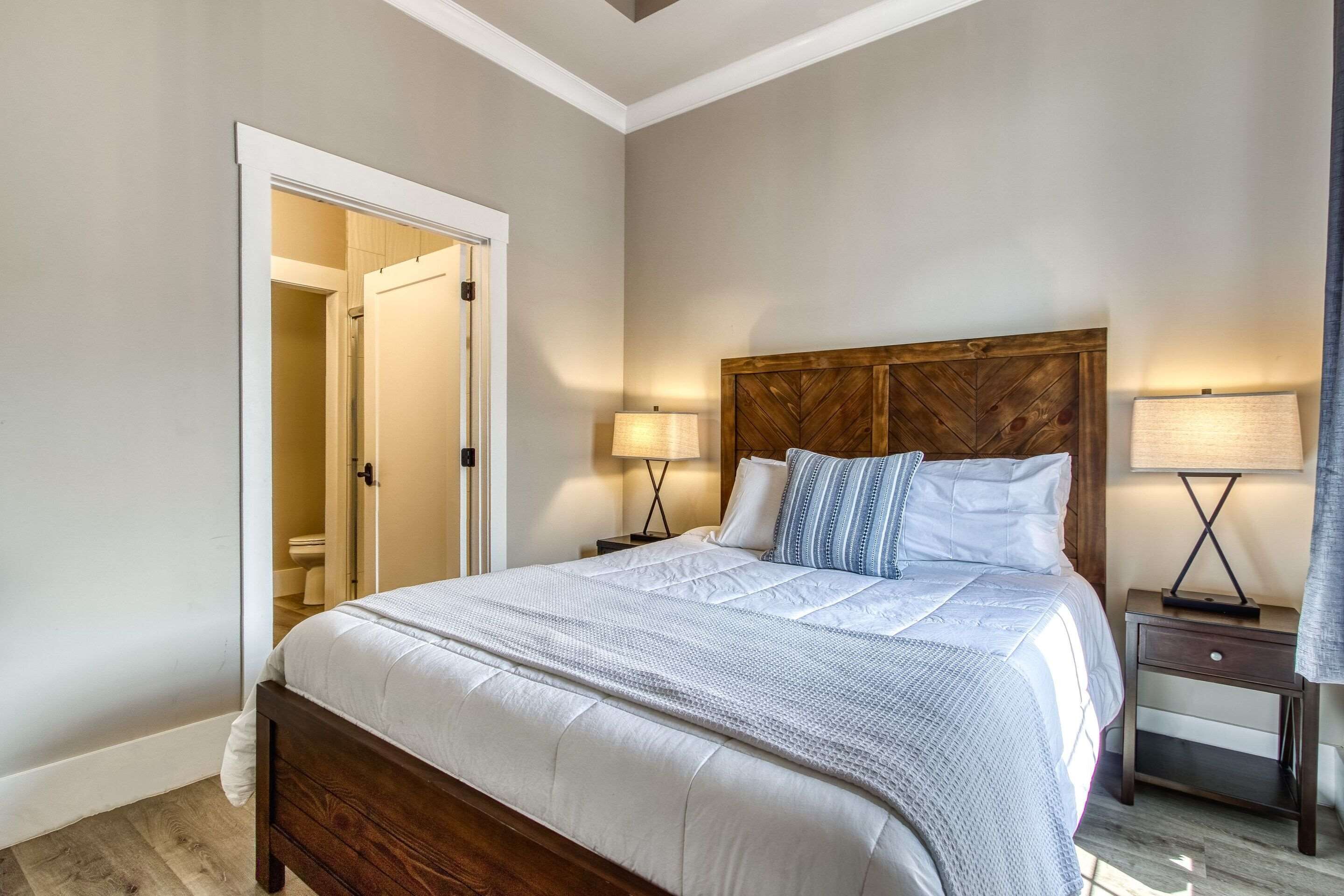 A bedroom with a large bed, wooden headboard, bedside lamps, and a door leading to a bathroom.