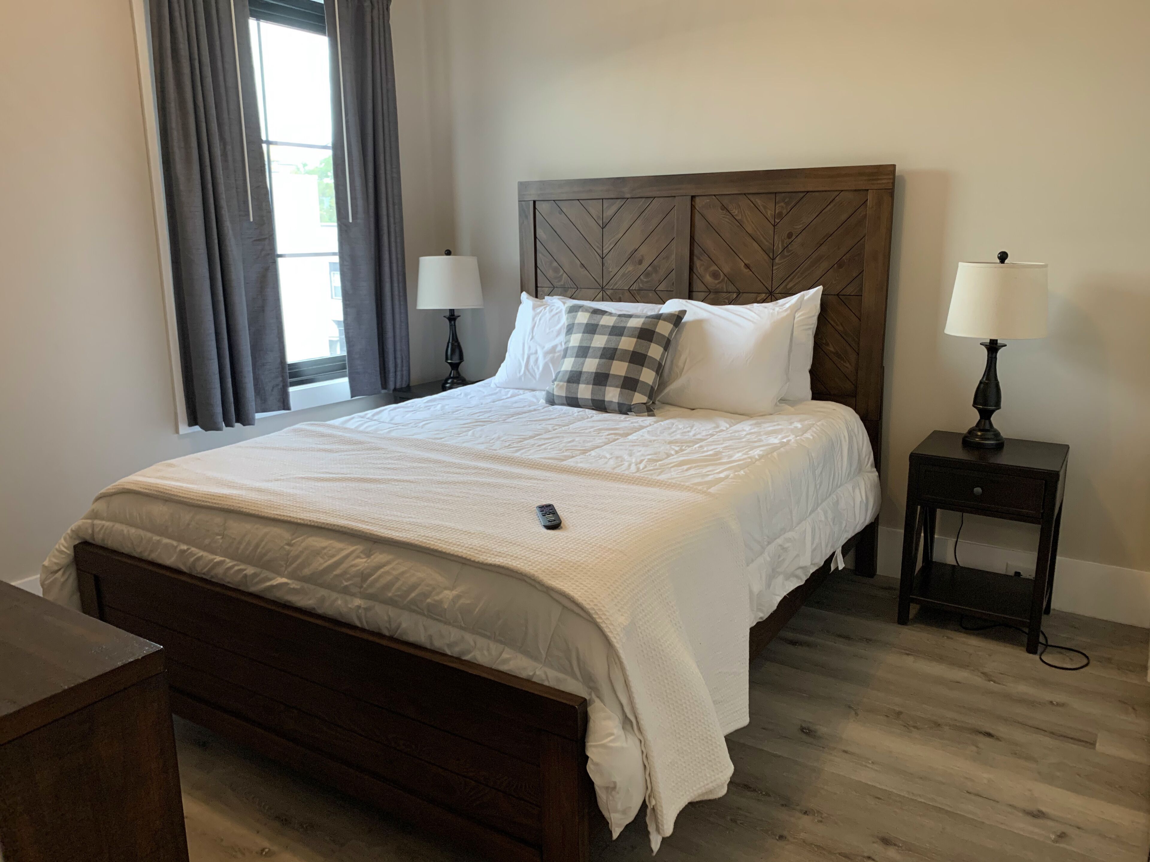Una cama bien tendida con cabecera de madera, dos lámparas de noche y una ventana con cortinas.