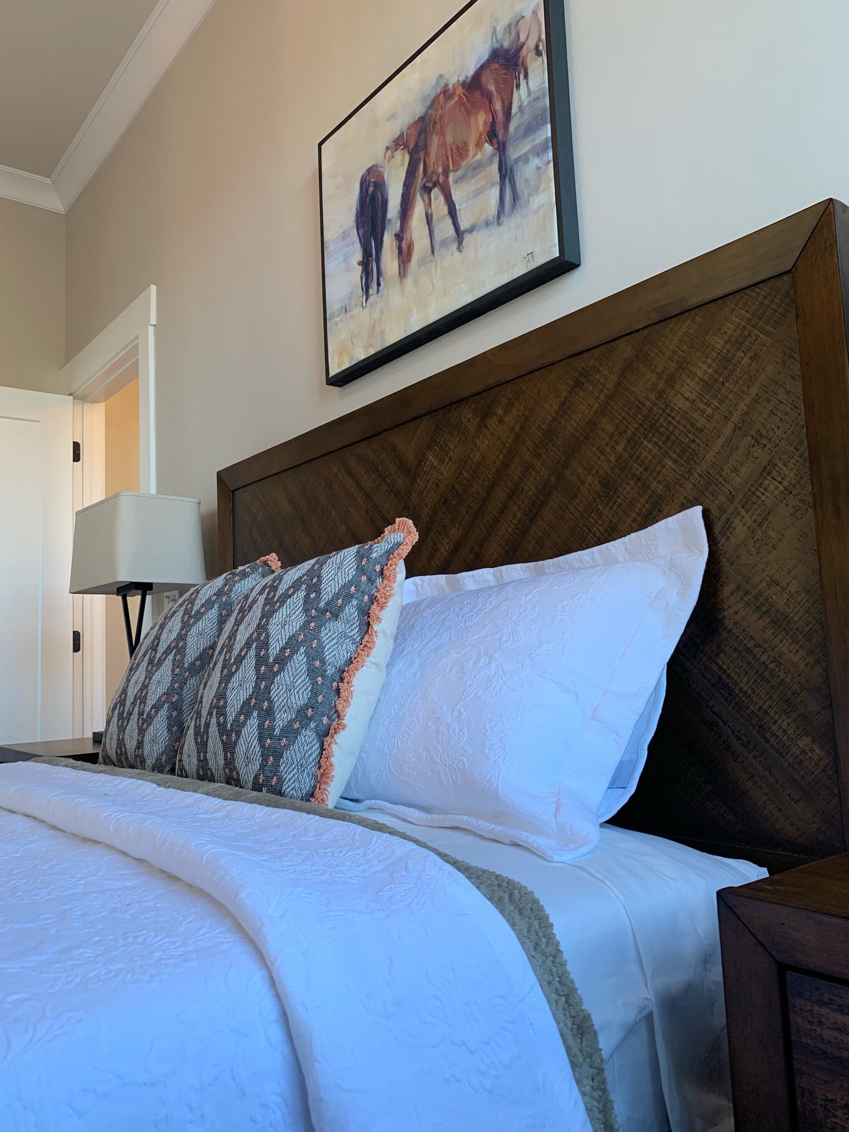 A neatly made bed with a patterned pillow and a plain pillow, a wooden headboard, and a framed painting of horses on the wall.