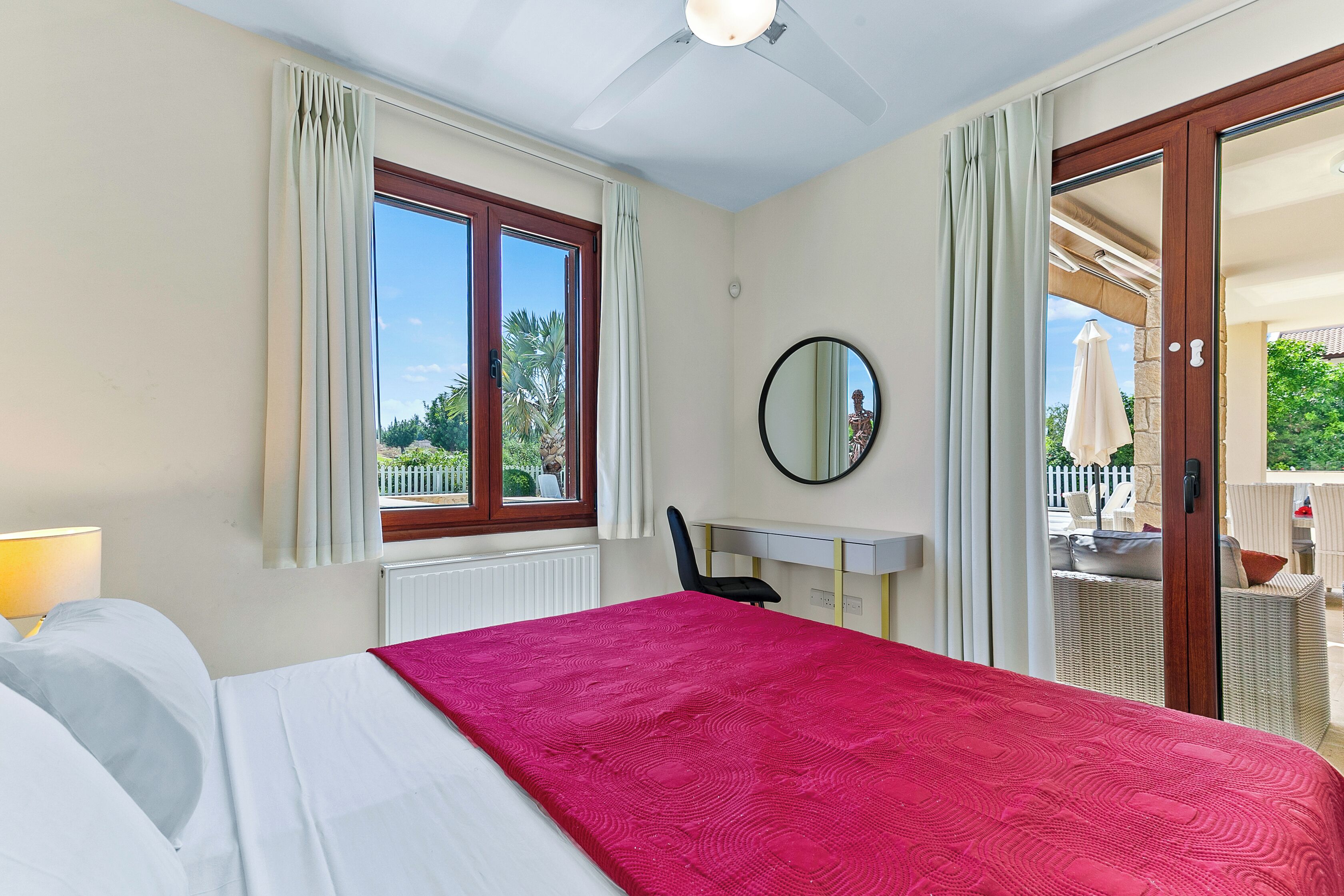 A hotel room with a bed, a desk, a chair, a window with curtains, a ceiling fan, and a view of the outside.
