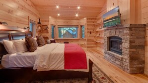 Un dormitorio rústico de cabaña con chimenea de piedra, cama con ropa de cama blanca y roja, y vista a una casa a través de una ventana.