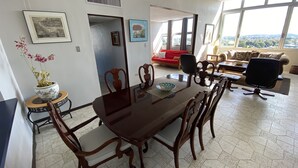 Een eetgedeelte met een grote houten tafel en stoelen, een rode bank en uitzicht door grote ramen.