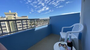Een balkon met een blauwe muur, een witte plastic stoel, een kleine ronde tafel en een fles wijn.
