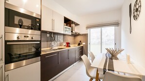 A modern kitchen with stainless steel appliances, a dining area with white chairs, and a large window providing natural light.