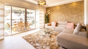 A modern living room with a beige sofa, a glass coffee table, and a large window with a view of buildings and greenery.