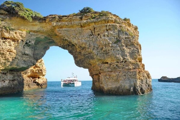 A natural rock arch formation with clear blue waters and a boat in the distance.