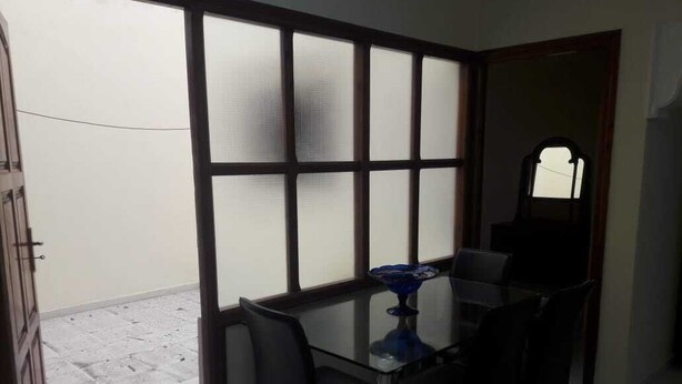 A room with a glass table and four chairs, a blue decorative item on the table, and a large window with frosted glass panels. - Furnished apartment in Essaouira (Essaouira)