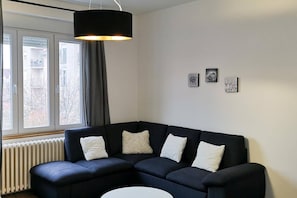 A modern living room with a dark blue sectional sofa, a white coffee table, and a black pendant light. There are two windows with curtains, a radiator, and three wall-mounted artworks. - Central 2 Bedroom Apartment 84 sq m with Private Parking (Novi Sad)