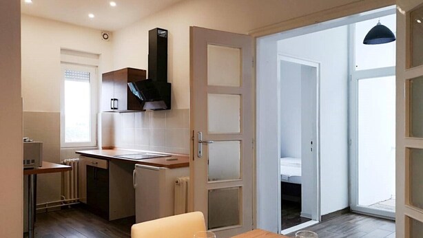 Cocina compacta con microondas, estufa y muebles de cocina modernos. Hay un comedor con una mesa dispuesta para dos personas, y se ve un dormitorio a través de una puerta abierta.