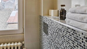 Un baño con salpicadero de azulejos, una ventana con vistas a edificios, un radiador y un perchero con toallas.