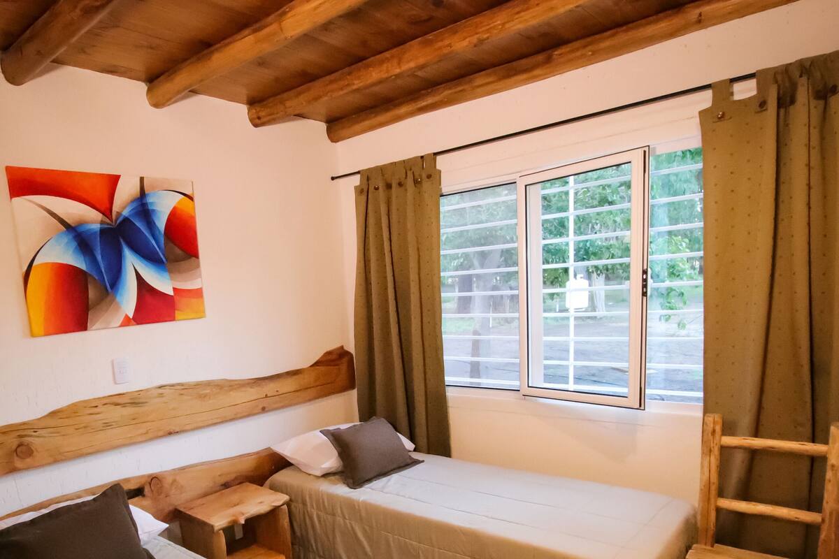 Una habitación pequeña y acogedora con cabecera de madera, una cama con almohadas, una mesita y una ventana con cortinas.