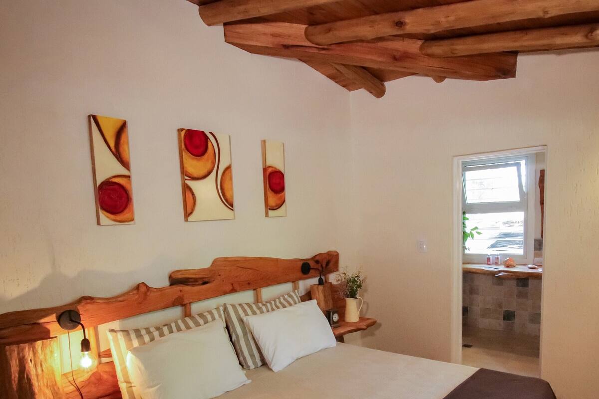 Un dormitorio con cabecera de madera, una cama con almohadas a rayas, una mesita de noche con lámpara y un televisor sobre un estante de madera.