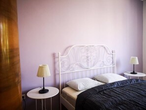 A bedroom with a white metal headboard, two bedside tables with lamps, a bed with a black cover, and a purple wall.