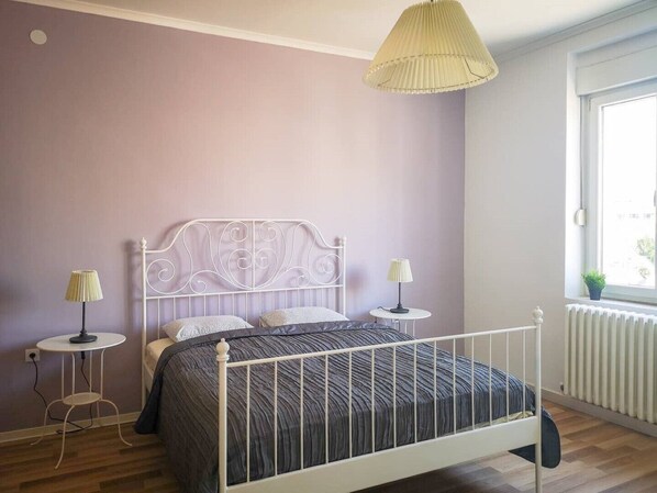 A bedroom with a white metal bed frame, a dark patterned bedspread, two bedside tables with lamps, and a window with a view of the outside.