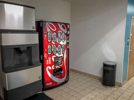 Máquina expendedora con varias opciones de bebidas, un basurero y una puerta en un pasillo.