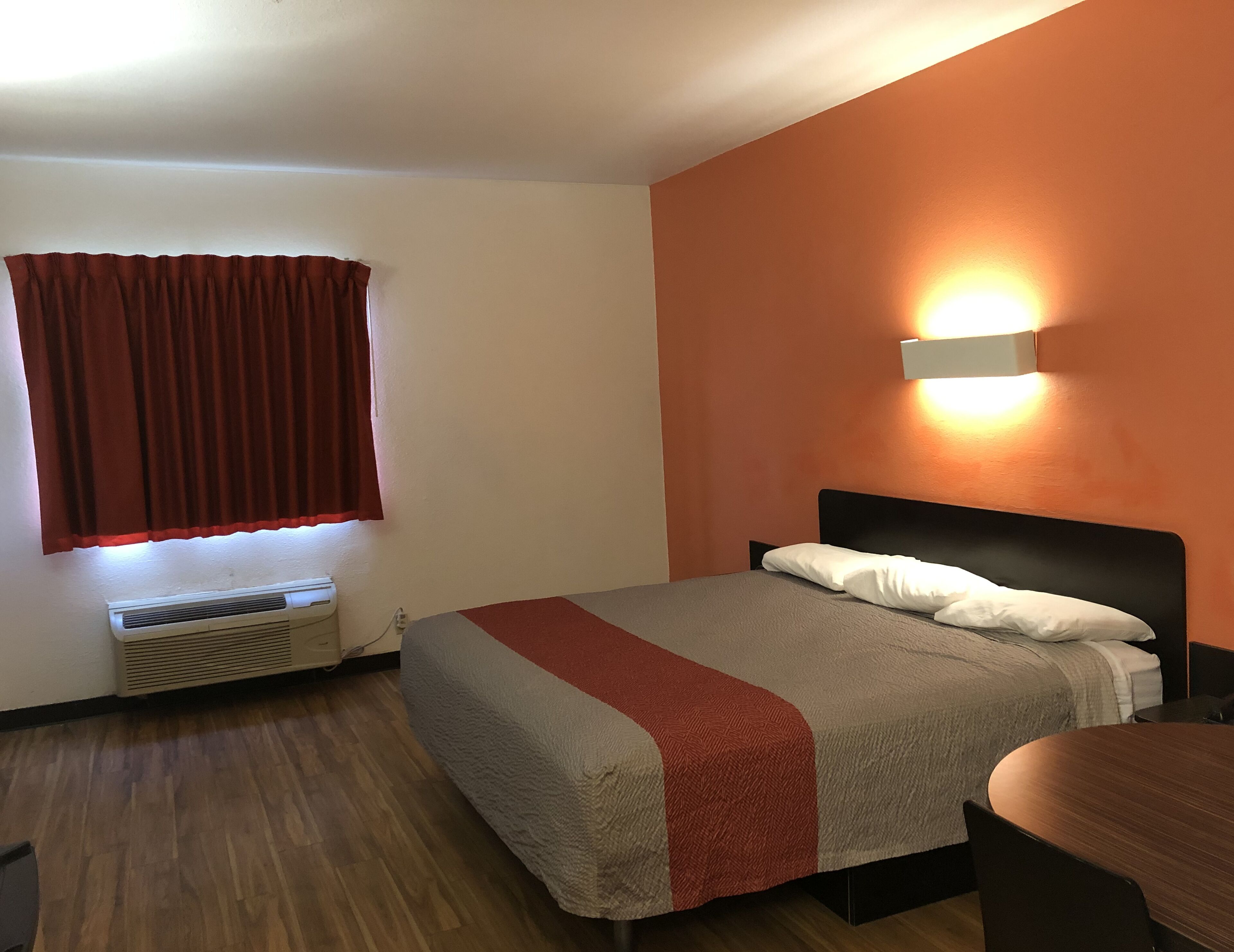 Une chambre d’hôtel avec un lit, une fenêtre avec des rideaux, un luminaire fixé au mur et un plancher en bois.