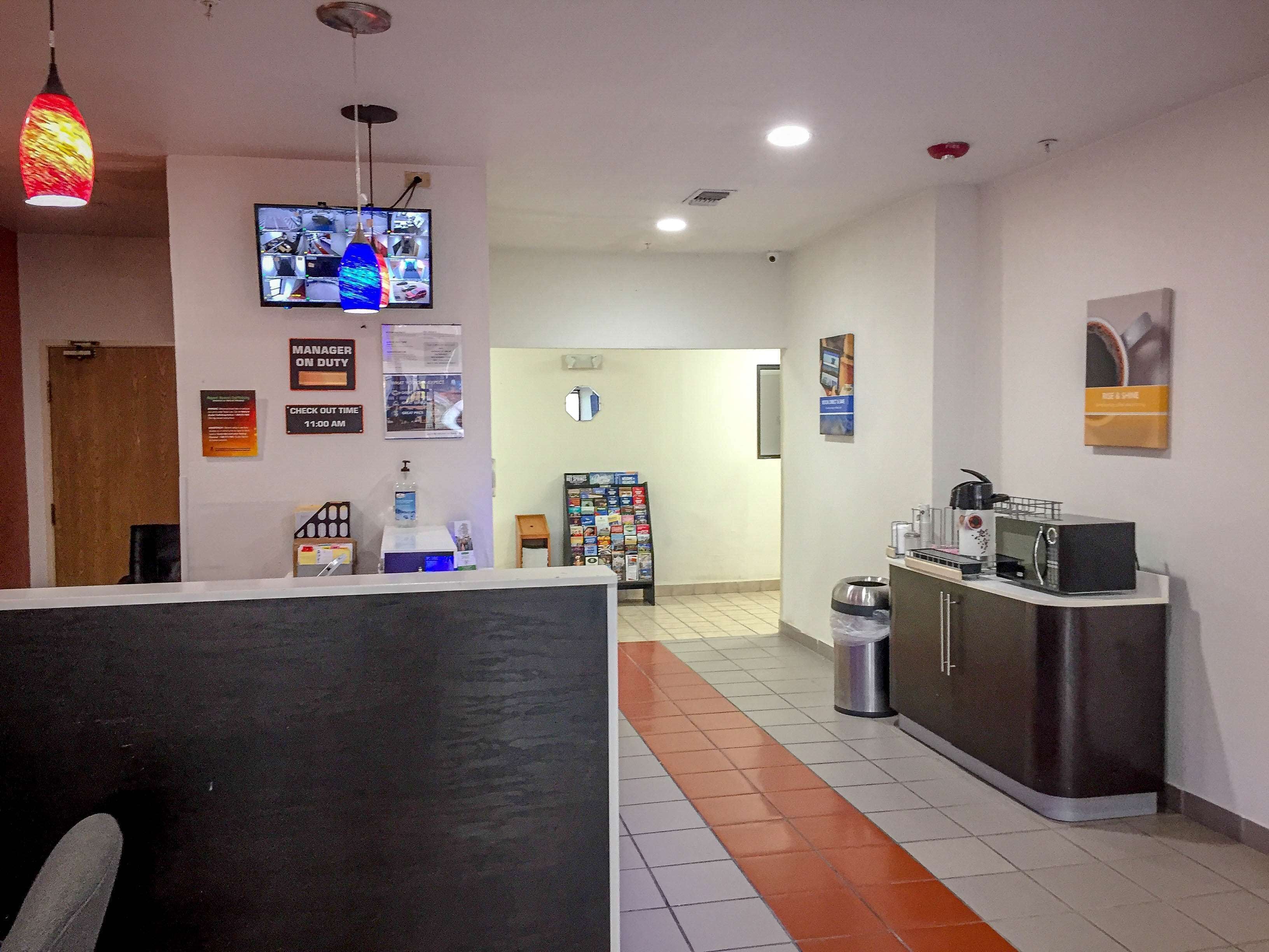 A hotel reception area with a check-in counter, a water dispenser, a waste bin, a refrigerator, and a television displaying various channels.