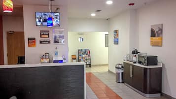 A hotel reception area with a check-in counter, a water dispenser, a waste bin, a refrigerator, and a television displaying various channels.