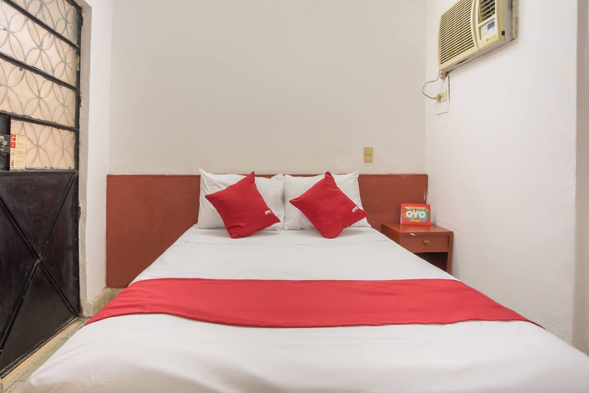 A hotel room with a bed, two red pillows, a red blanket, a nightstand with a card, and a window with a metal grill.