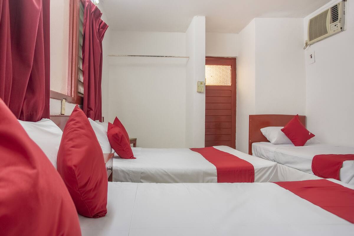 A hotel room with three single beds, each with red bedding and pillows. There is a wooden headboard on the right bed, and a small wooden table between the beds. The room has white walls and a wooden door with a window above it.
