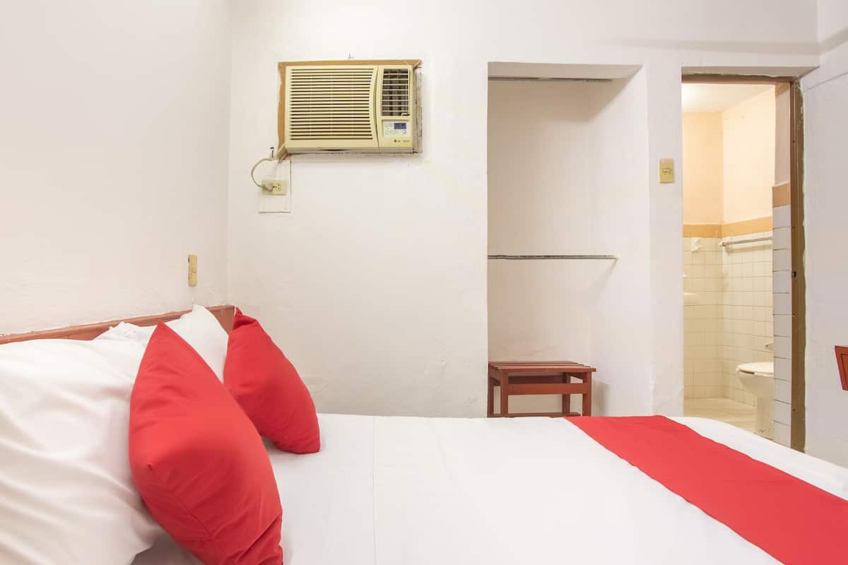A hotel room with a bed, red and white pillows, a small wooden table, a wall-mounted air conditioner, and a bathroom visible through an open door.