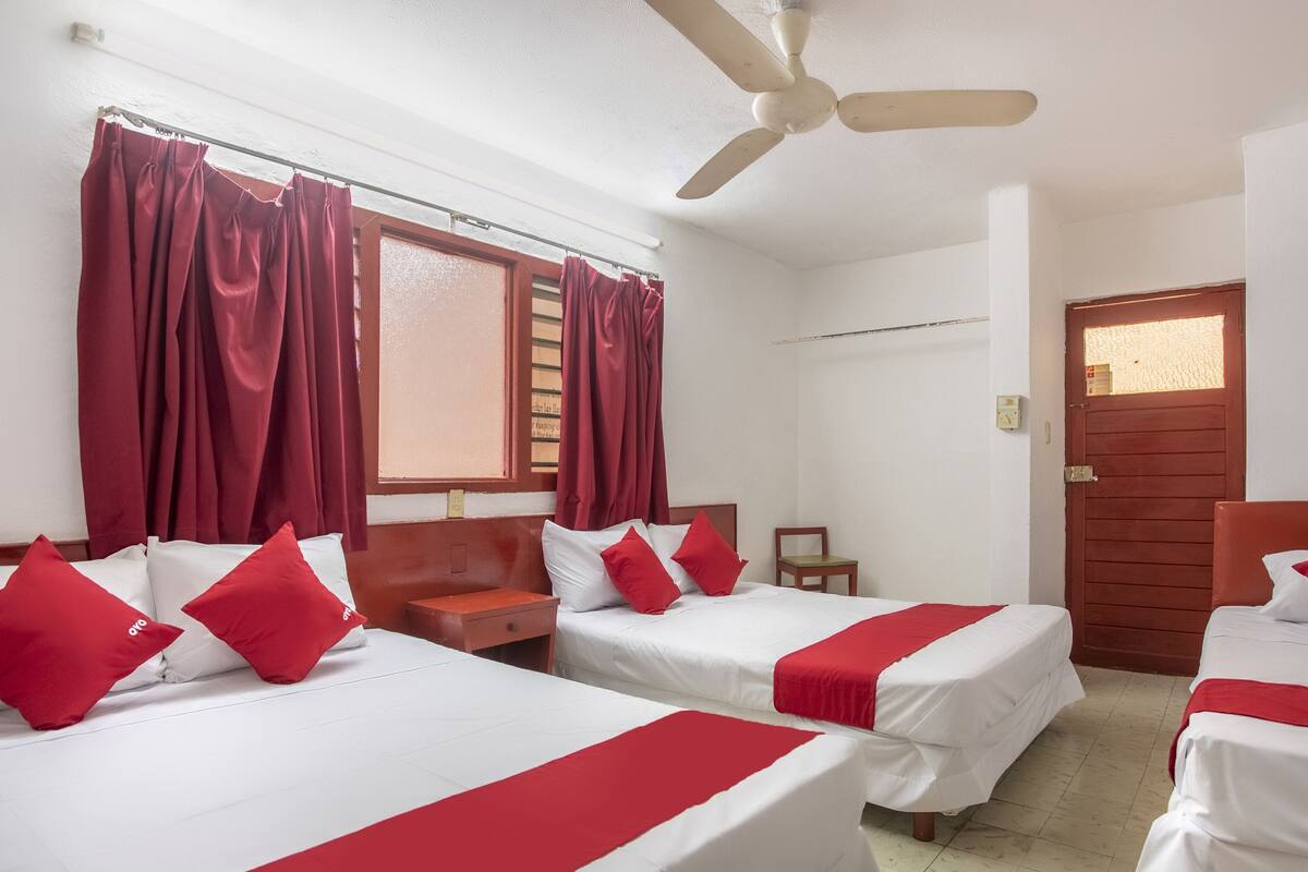 A hotel room with four beds, a ceiling fan, a window with curtains, a wooden door, and a small wooden chair.