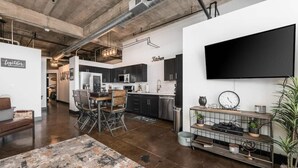 Una cocina moderna con gabinetes oscuros, un comedor con mesa y sillas de madera, y una sala de estar con televisor y planta.