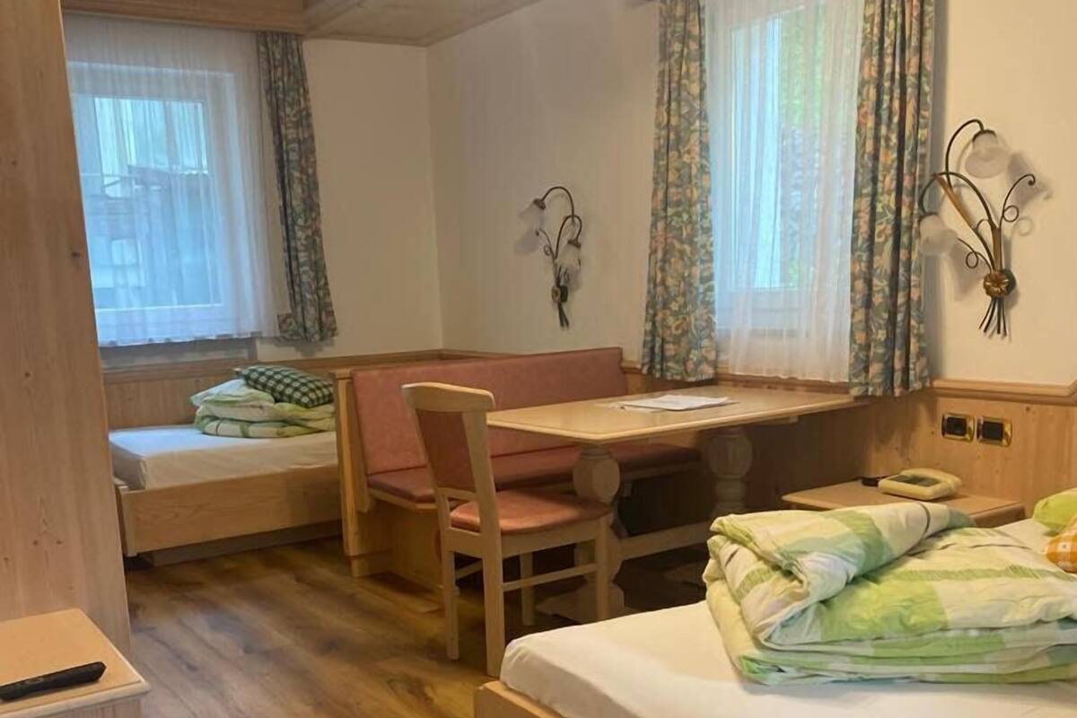 Ein gemütliches Zimmer mit einem Bett, einem Schreibtisch und einem Sessel. Ein Fenster mit Vorhängen und eine Wandleuchte sind vorhanden.