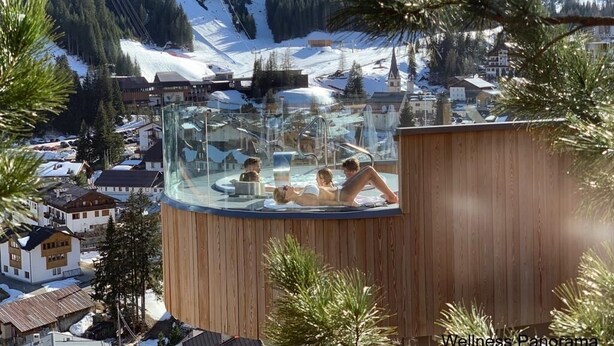 A glass-enclosed hot tub with a wooden exterior, surrounded by snow-covered mountains and pine trees. - Hotel Olympia (Livinallongo del Col di Lana)