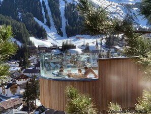 Un bain à remous en verre, doté d’une structure extérieure en bois, entouré de montagnes enneigées et de pins.