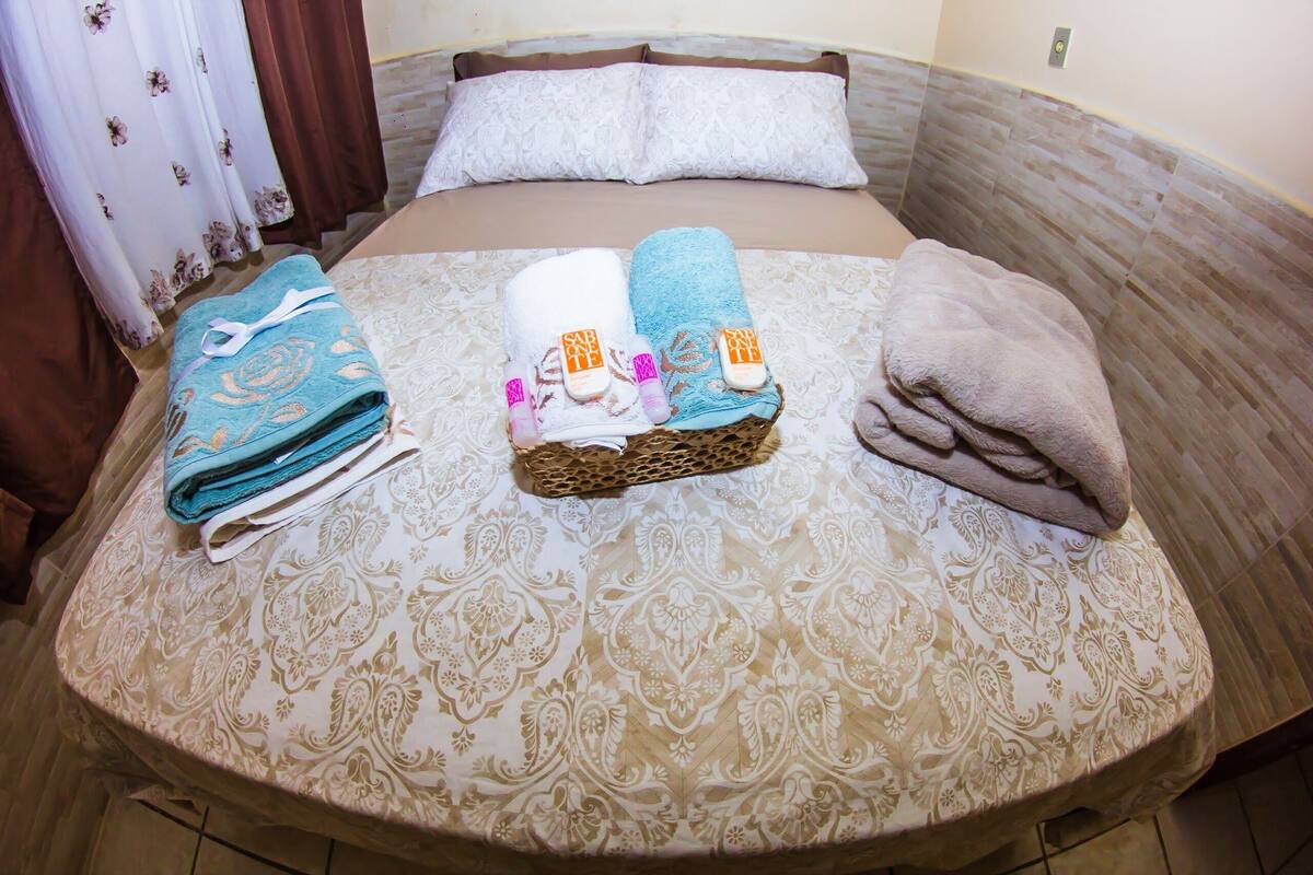A neatly made bed with a patterned bedspread, two folded towels, and a rolled-up blanket.