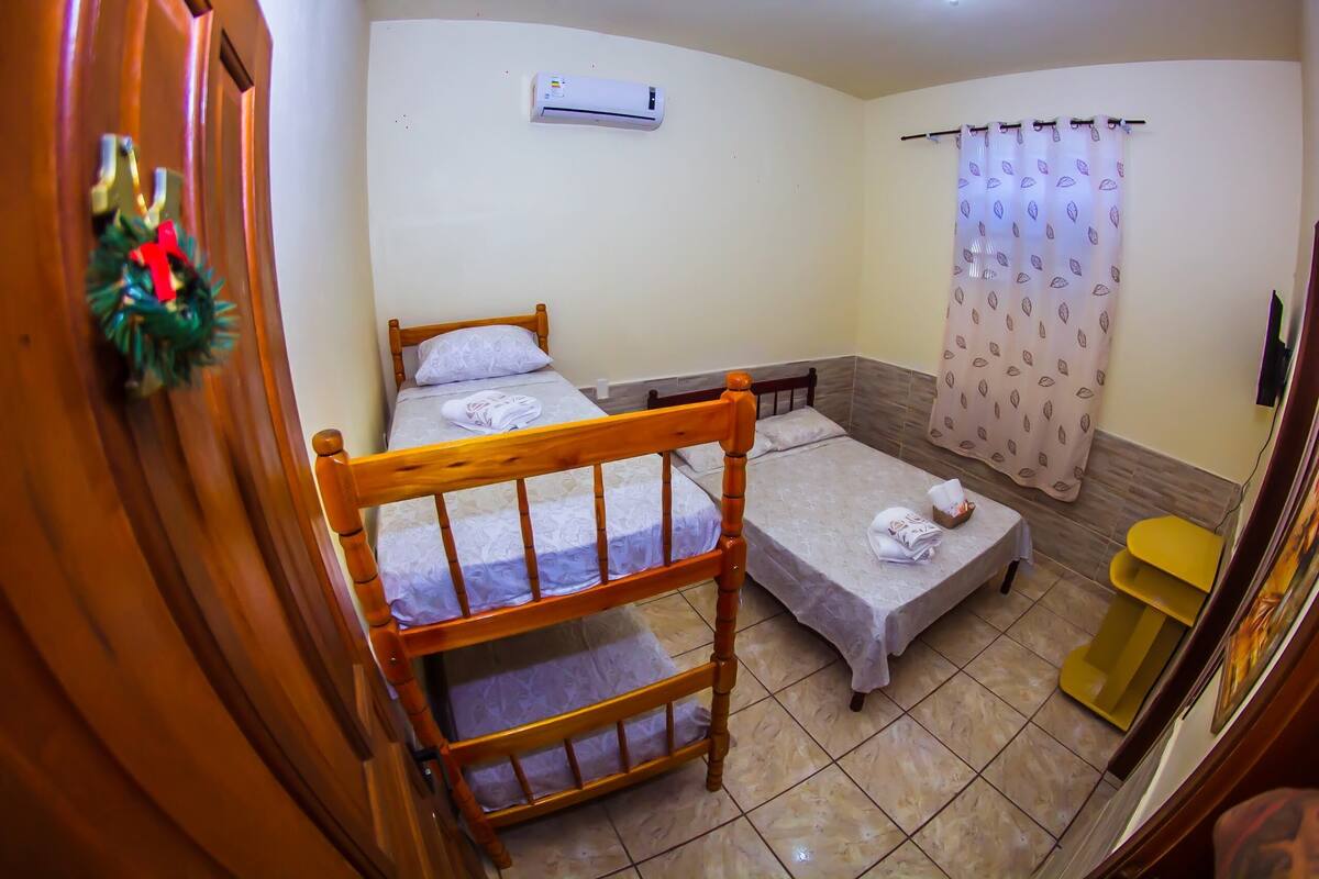 A room with two beds, a wooden headboard, a small table, a yellow side table, a curtain, and an air conditioning unit.