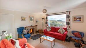 A living room with a fireplace, a red sofa, a blue armchair, and a view of the countryside.