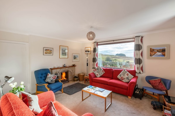 A living room with a fireplace, a red sofa, a blue armchair, and a view of the countryside. - Laghead Cottage -  a cottage that sleeps 6 guests  in 3 bedrooms (Gatehouse of Fleet)