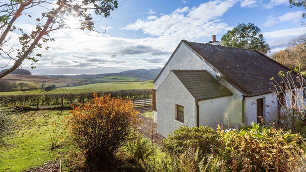 A small white house with a sloped roof, surrounded by lush greenery and a scenic view of rolling hills.