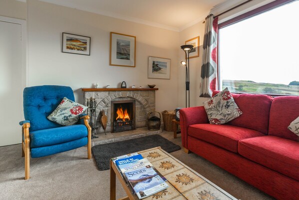 A living room with a fireplace, two sofas, a coffee table with magazines, and a view through a window.