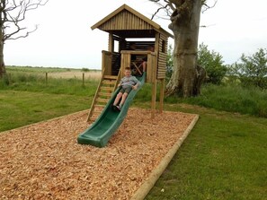 Explore the fun of a wooden play structure with a green slide, ladder, and upper platform, set amidst a grassy area surrounded by trees.