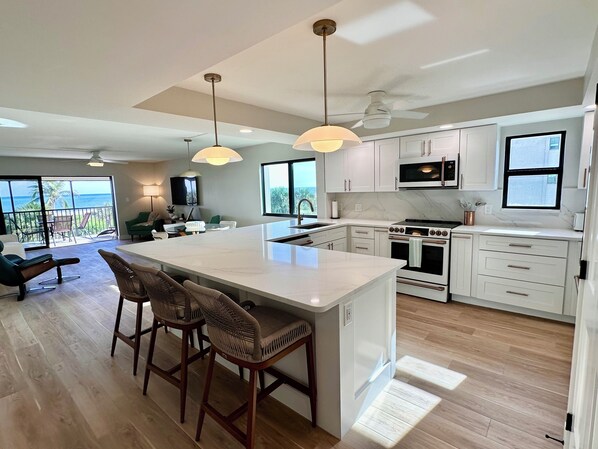 A modern kitchen with white cabinets, a large island with bar stools, a built-in oven, and a view of the ocean through large windows.