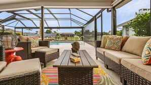 Un espace extérieur aménagé, abrité et doté de mobilier en osier, d’une table en bois, et offrant une vue sur une piscine et des maisons en arrière-plan.