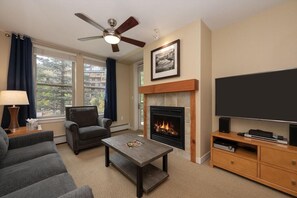 A living room with a fireplace, a flat-screen TV, a ceiling fan, and a sofa. - Fraser Crossing 3227 Ski In Ski Out Winter Park (Winter Park)