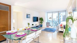 A modern dining area with a glass table, white chairs, and a city view through large windows.