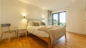 Un dormitorio con piso de madera, una cama con cabecera, mesitas de noche, una silla y una ventana con vistas.