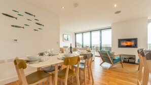 Un comedor moderno con una mesa de madera dispuesta para cuatro personas, rodeada de sillas con cojines verdes. Se observa una chimenea al fondo, y un ventanal ofrece vistas al exterior.