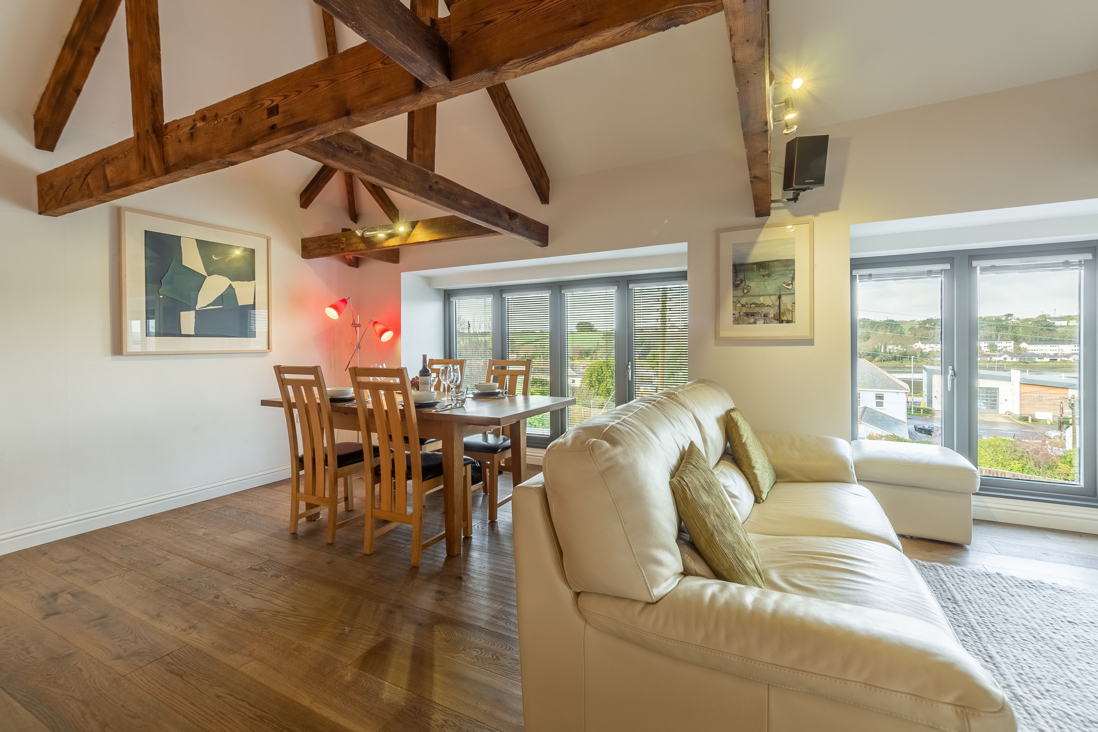 A spacious living area with a wooden ceiling, a dining table with chairs, a white sofa, and large windows offering a view of the outside.