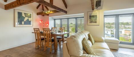 A spacious living area with a wooden ceiling, a dining table with chairs, a white sofa, and large windows offering a view of the outside.